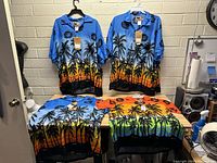 Four new Hawaiian shirts displayed on hangers and folded on a table, showing vibrant tropical palm tree and sunset designs in blue, orange, green, and black colors with tags attached.