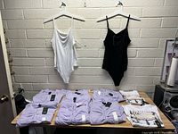 Two sleeveless bodysuits (white and black) hanging on a wall with multiple pairs of light purple pants and folded patterned tops on a table under them