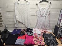 Photo shows 11 maternity clothing items laid flat including tops and dresses in various colors and patterns; white lace long sleeve top, white sleeveless dress with print, floral and abstract patterned tops, and casual tops in pink, purple and black.
