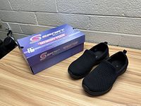 Pair of black Skechers women's slip-on shoes next to their box on a wooden surface with visible brick wall background.
