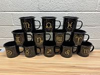 Front view of all 12 black enamel mugs showing all zodiac signs in a stacked pyramid arrangement on a wooden surface with a white cinder block wall background.