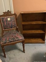 Full view of the antique wooden chair with patterned upholstery and wooden bookcase with three shelves side by side.