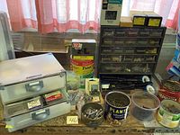 Overall view of assorted fasteners in metal drawers, open tins, plastic containers, and labeled packaging showing variety of screws, nails, hooks, and small hardware pieces.