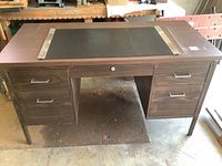 Full front view of the wood-finish office desk showing the black writing surface with metal trim and the drawer configuration.