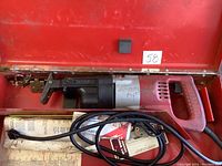 Milwaukee Sawzall power tool inside metal case, showing side view and red handle grip, with power cord coiled