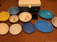 Photo showing an assortment of plastic and glass serving bowls and trays in various colors and designs, alongside an Igloo cooler.