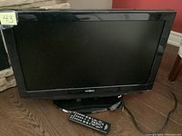 Front view of the 26-inch Insignia LCD TV positioned on a wooden floor with remote and cords visible.