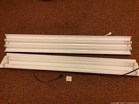 Photo showing two white fluorescent light fixtures with three tubes each, laid on a carpeted floor, with one fixture showing its hanging chain and cord with plug.