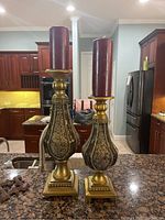 Two tall ornate metal candlesticks with mosaic inlay bases holding tall burgundy pillar candles photographed on kitchen counter.