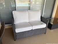 Outdoor faux wicker loveseat with white cushions shown with a patio background and sliding glass doors.