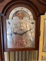 Close-up of clock face showing intricate silver dial with gold Arabic numerals, decorative black hands, and moon phase dial at top.