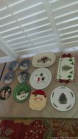 Full view of all holiday plates arranged on beige fabric, showing different designs including Santa Claus, trees, holly, nutcracker, and candy cane border