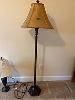 Full view of the floor lamp with beige fabric lampshade next to wall outlet and part of table lamp visible.