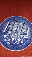 Close-up photo of a single 2011 Royal Copenhagen Christmas porcelain plate showing detailed scene of a child decorating a Christmas tree with lit candles, bordered by pine and star motifs.