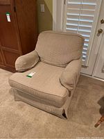 Front angled view of beige upholstered armchair showing cushioned backrest, seat, and padded armrests with skirted bottom on carpeted floor.