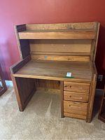 Wood desk with three drawers on the right side, visible scratches, and an unattached top shelf, photographed against a red wall on carpet.
