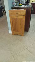 Front view of wooden garbage pail cabinet with drawer and door panel closed, showing two decorative tiles on the top.