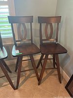 Two matching wooden swivel stools with dark finish and elliptical cutout design on backrest, angled legs on tiled floor near window.