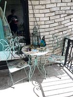 Photo showing round bistro table and two butterfly-back chairs with floral and butterfly painted designs on a tiled outdoor floor near a brick wall and glass door.