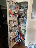 Closet with door-mounted wire rack filled with small cleaning bottles and household products, mop or broom leaning inside.