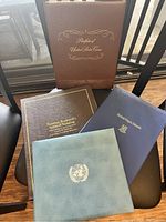 Four books stacked showing covers: burgundy US coin binder, brown Norman Rockwell book, navy British Virgin Islands book, light blue United Nations binder