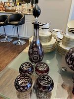 Tall cranberry colored etched glass decanter with stopper on a glass surface alongside four round cranberry glass cups, all decorated with detailed etched floral and grape designs.