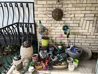 A wide view showing an assortment of outdoor gardening items including pots, decorative metal sun wall art, colorful flower stakes, watering cans, and gardening tools on a porch.