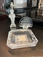 Three Lalique crystal items displayed on black surface: frosted male figurine, ship paperweight, and square bowl