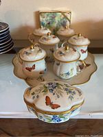 Photo showing 6 lidded demitasse cups arranged on a scalloped tray and a matching covered decorative box with butterfly and floral designs and gold trim.