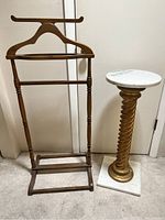 Wood vallet stand and marble top pedestal table side by side on carpeted floor.