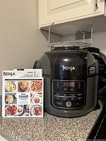 Front view of Ninja Foodi pressure cooker with control panel and user manual placed next to it on countertop.