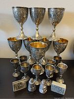 Wide view of the full lot showing 12 ornate wine goblets (6 large, 6 small), 2 match holders, and 2 salt and pepper shakers arranged on a table