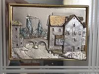 Close up front view of the 3D silver relief sculpture depicting houses, bridge, trees, and swans on water