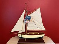 Front-left angled view of 1930s style yacht model showing sails, rigging, and wooden base with certificate tag attached.
