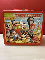 Front view of vintage Disney Express metal lunch box featuring multiple classic Disney characters on a train at Station #1. Bright red handle and edges.
