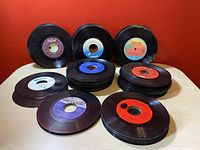 Stacks and scattered 45 RPM vinyl records arranged on a light wooden surface with orange background wall, showing various label colors and wear.