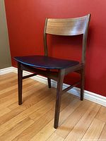 Front angle view of the teak chair with black seat, showcasing the wood grain and curved backrest against a red wall and hardwood floor.