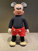 Front view of vintage Marching Mickey doll showing red shorts with white buttons, black fabric top, yellow shoes, and molded plastic head and hands.