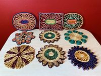 Top view of nine colourful wicker flower and rectangular trivets arranged in three rows on a light-colored surface with a red wall background.