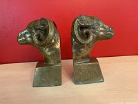 Pair of brass ram head bookends photographed from the left side, showing the detailed horns and patina on the surface.