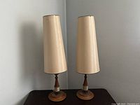 Front view of both matched table lamps placed side by side on a dark wood surface, showing tall beige shades and teak and ceramic bases.