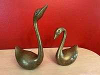 Pair of brass swan figurines on a light wooden surface against red background showing side and front views with natural tarnished patina.
