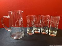 Clear glass pitcher with etched floral design and four matching glasses with similar floral etching, photographed against a red background for visibility.