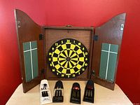 Front view of open wooden dart board cabinet showing dart board inside and chalk scoreboards on inner door panels with 4 sets of darts in front.