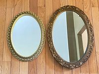 Two vintage oval wall mirrors on wood floor: one with ornate carved wood frame, the other with gold-tone plastic frame with bead details.