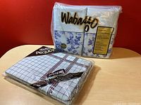 Wabasso double sheet set in sealed packaging next to Pequot flat sheet in sealed packaging on wooden table against red wall.