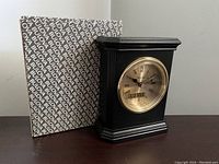 Black vintage Bulova mantle clock with gold-tone bezel, original box with Bulova branding, and instruction paper laid out on table.