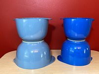 Two sets of vintage Danish Rosti mixing bowls in light and bright blue colors stacked on top of each other on a table against a red wall background.
