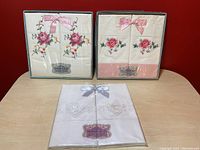 Three sets of vintage embroidered pillow cases displayed on a table, showing original gift boxes and packaging with satin bows and floral embroidery.