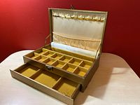 Vintage golden jewelry box opened showing small square compartments inside and slide out drawer below on beige wooden surface against red background.